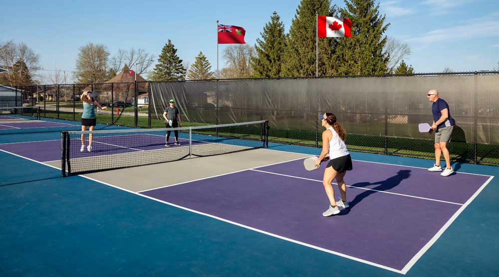 Four players engaged in a competitive pickleball doubles match on an outdoor court in Canada, with the Ontario and Canadian flags flying in the background.